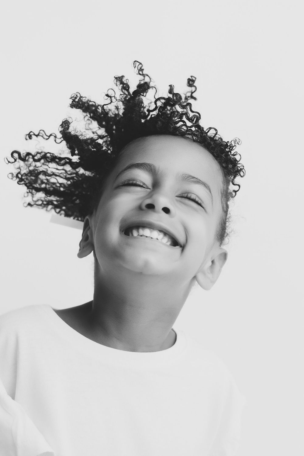 Black and white portrait of a joyful child with curly hair, eyes closed, smiling brightly against a light background.