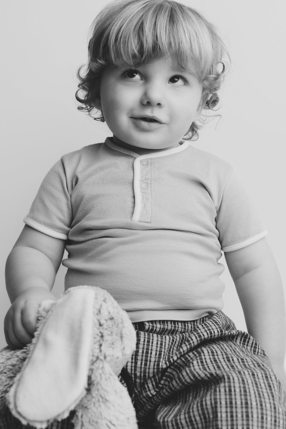 Black and white photo of a cute toddler with curly blonde hair sitting with a stuffed animal toy.