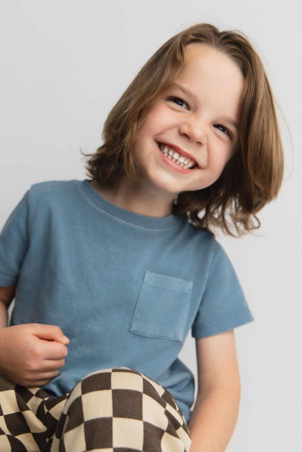 Happy young child smiling brightly, wearing a blue t-shirt and checkered pants against a white background.
