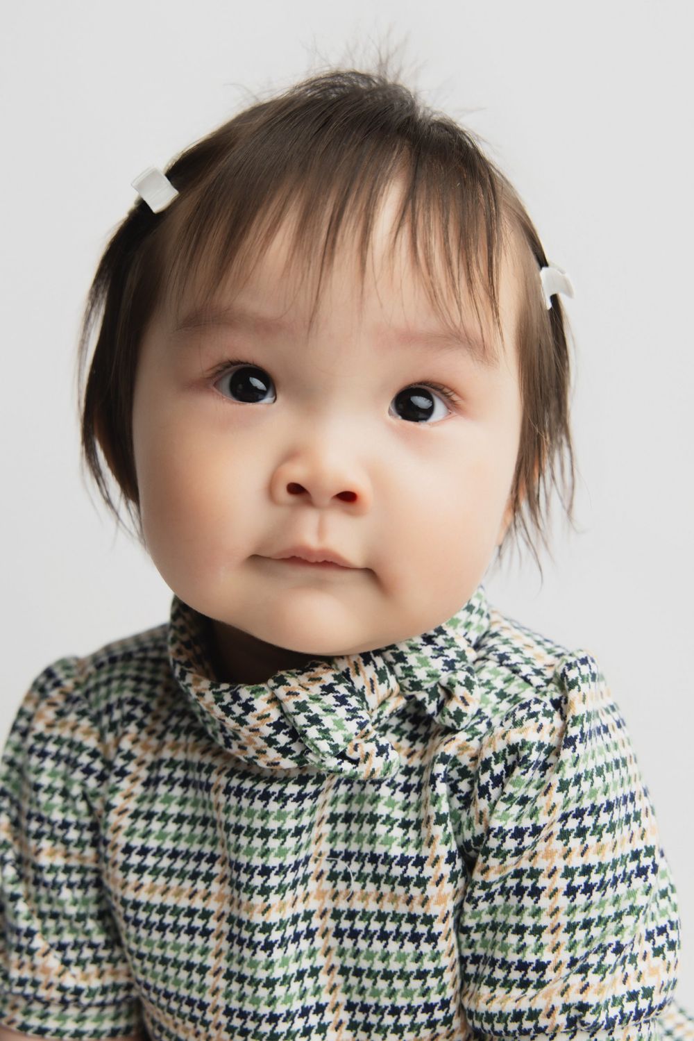 Adorable Asian baby girl wearing a houndstooth dress with white hair clips, looking curiously at the camera.