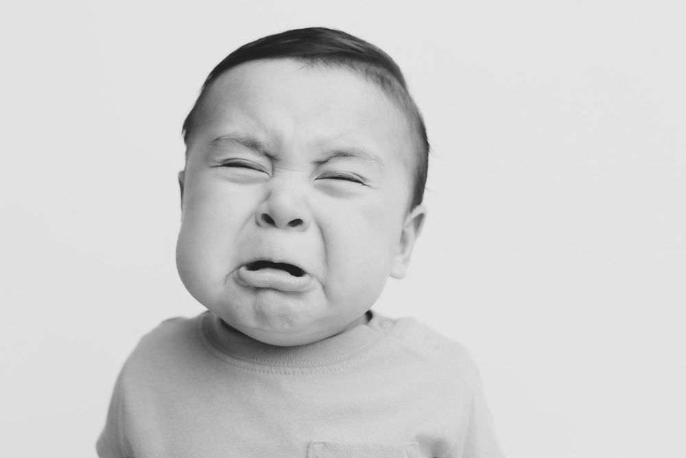 Black and white photo of a crying newborn baby with scrunched face and closed eyes wearing a light onesie.