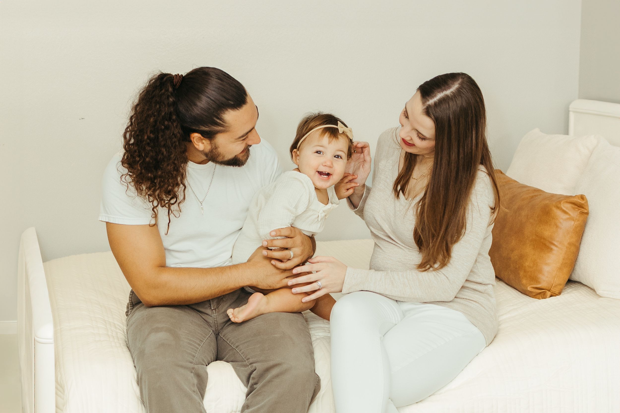 Young family with curly haired dad and mom smiling with happy baby on white couch in Pearland