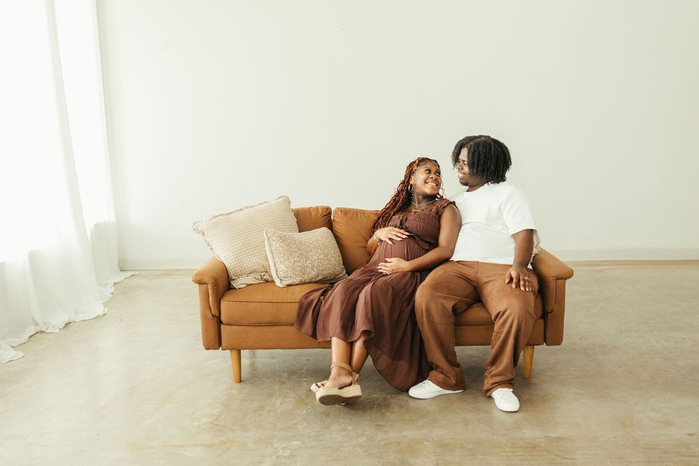 Pregnant woman in brown dress sitting on leather sofa with partner during maternity session.