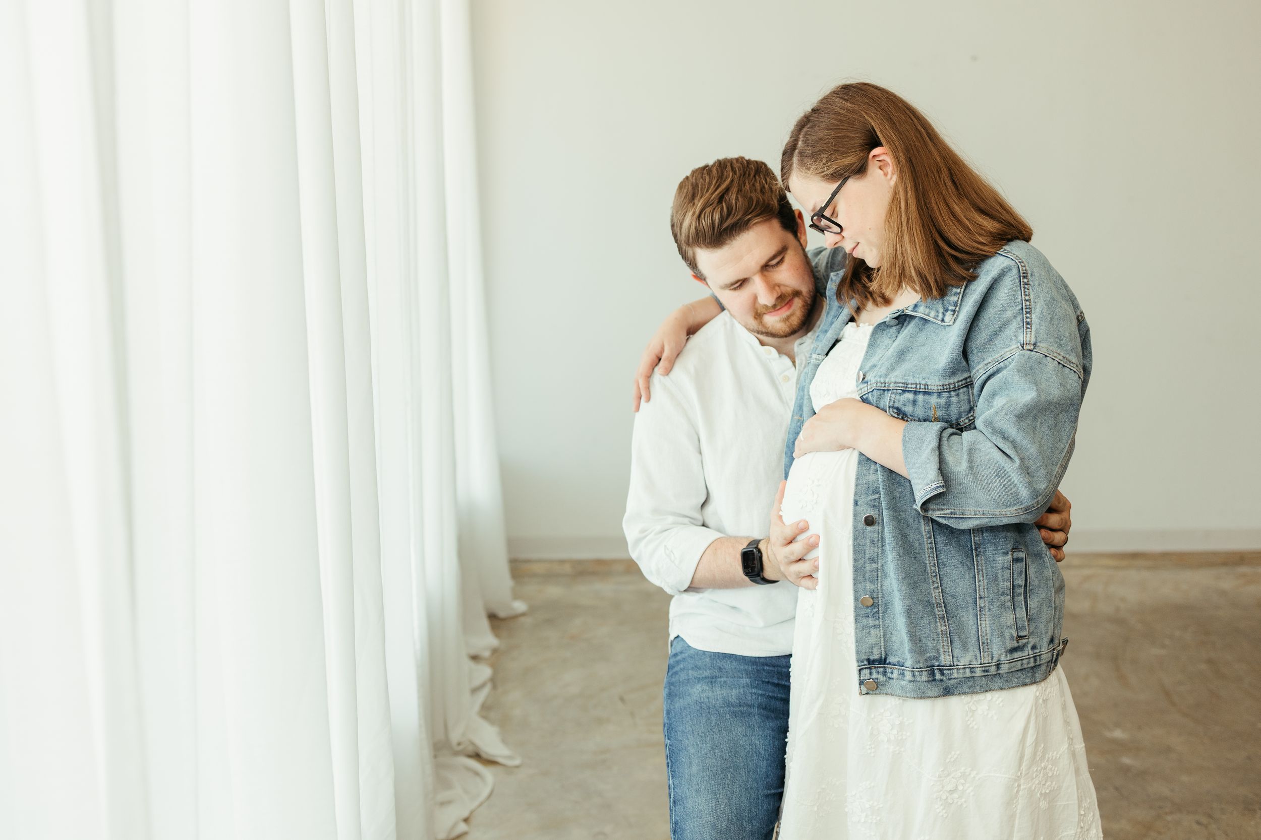 Expectant couple embracing tenderly near white curtains during indoor maternity photo session in Pearland Texas