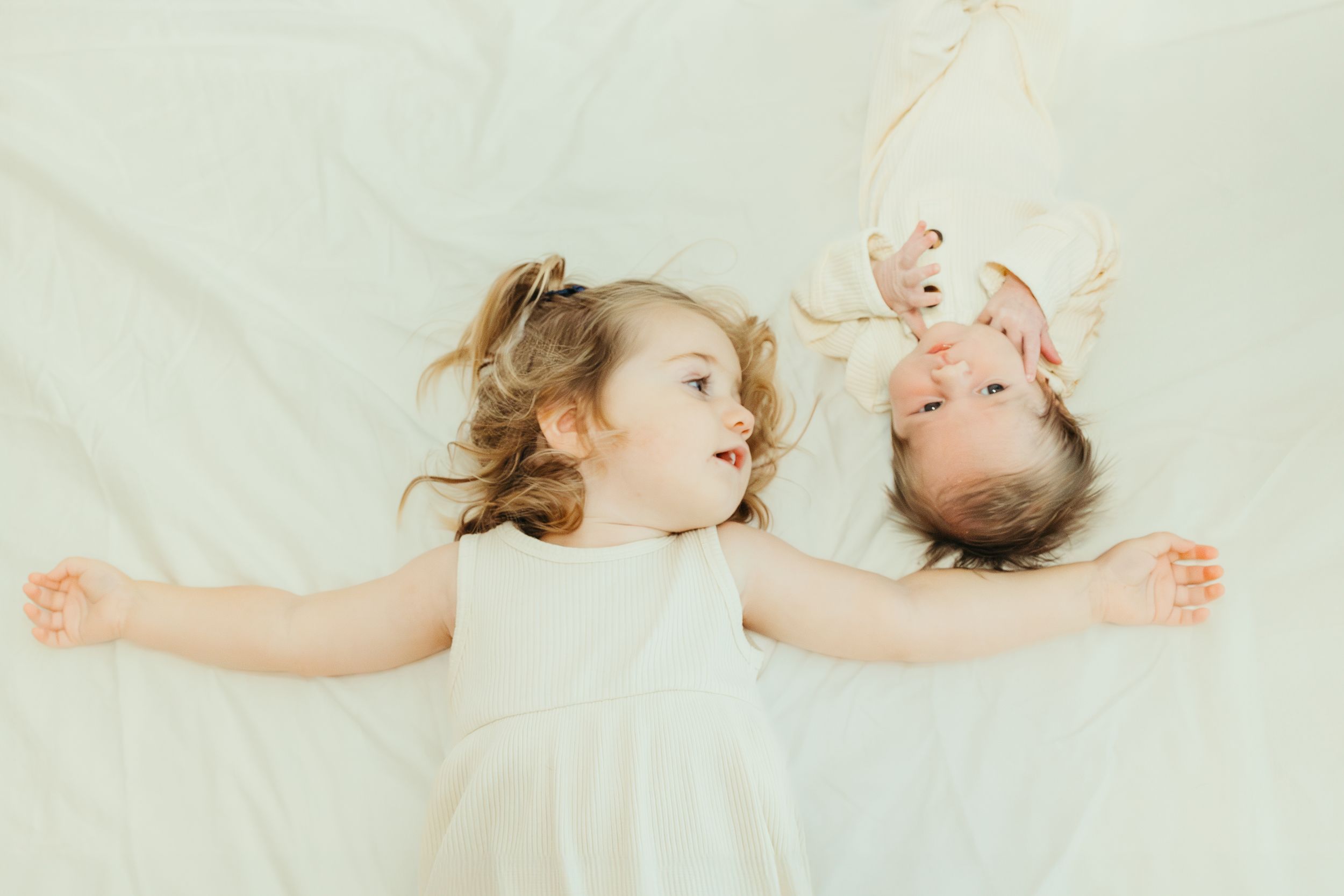 Two young children in white dresses lying on white bedding with arms outstretched in Pearland Texas