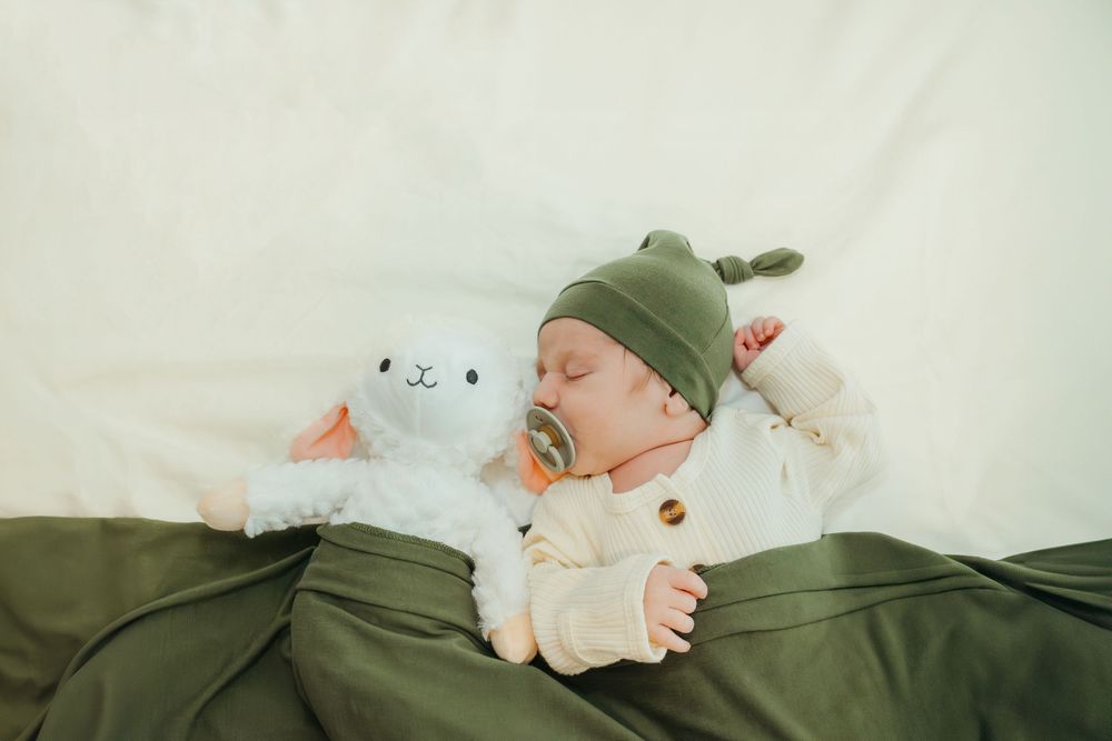 Sleeping newborn baby in green hat cuddling a white lamb stuffed animal on green blanket.