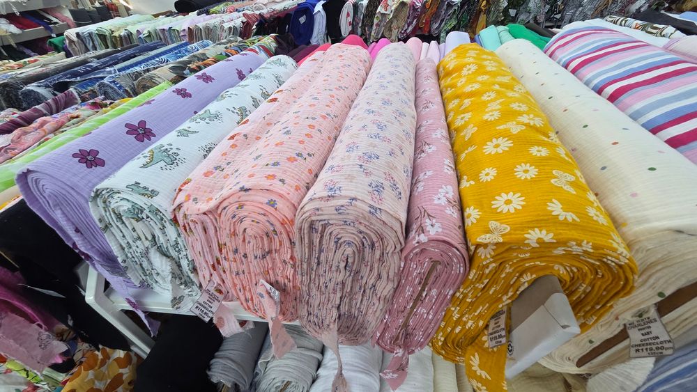 Colorful rolls of patterned fabric displayed on a market stall, including floral and printed textiles in pink, yellow, and purple.