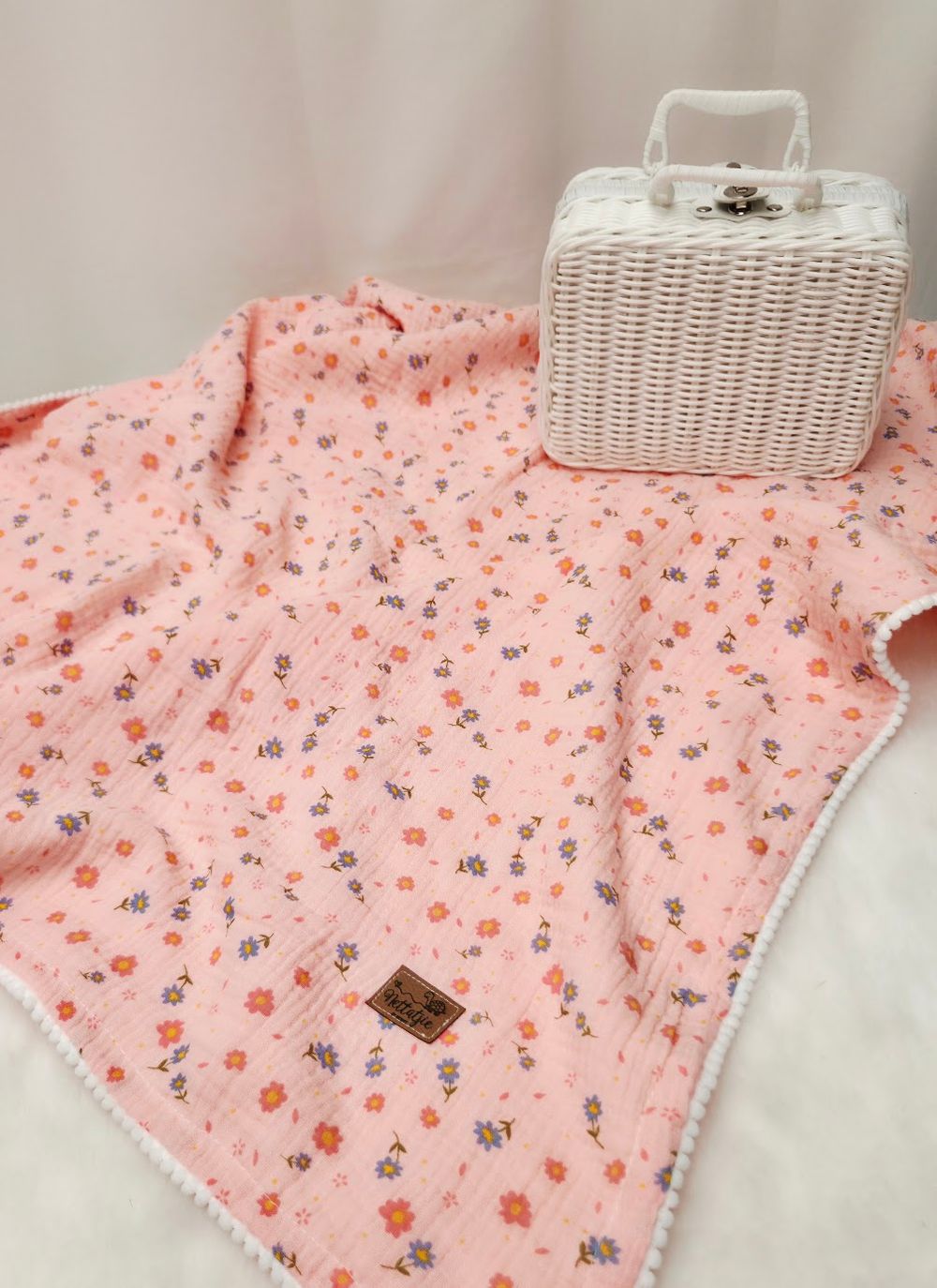 Pink floral muslin baby blanket with white pompom trim displayed next to a white wicker basket.