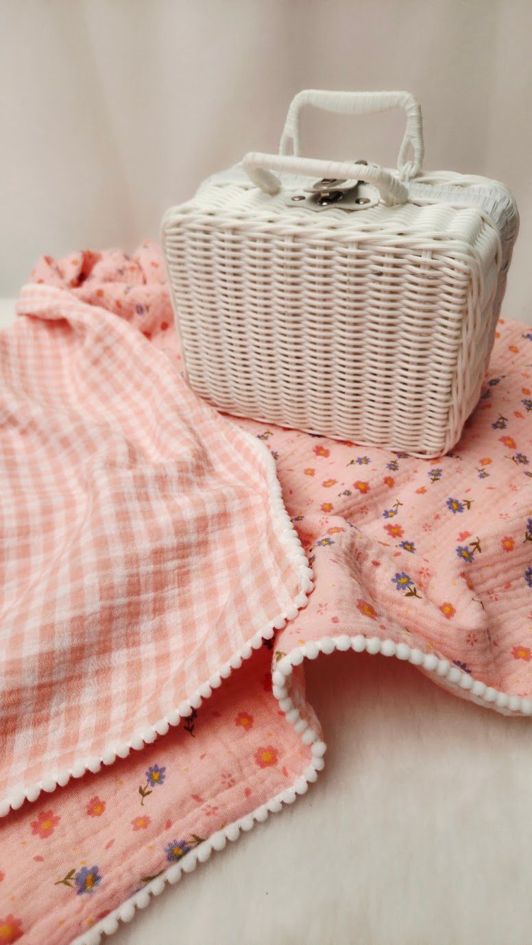 Pink gingham and floral muslin blankets with pom-pom trim beside a white wicker basket purse.