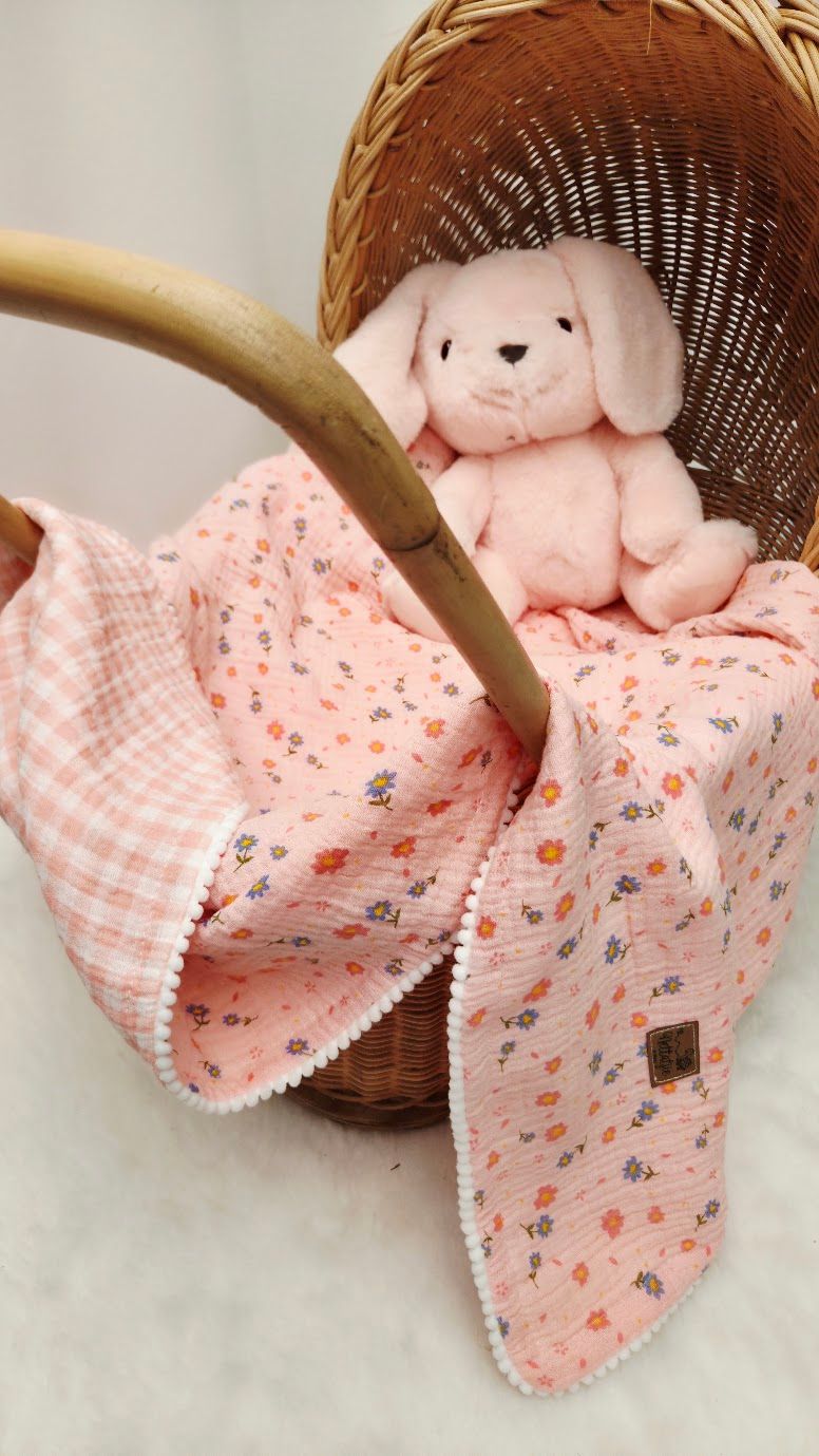 Pink floral muslin baby blanket with gingham trim and bunny stuffed animal in a wicker basket.