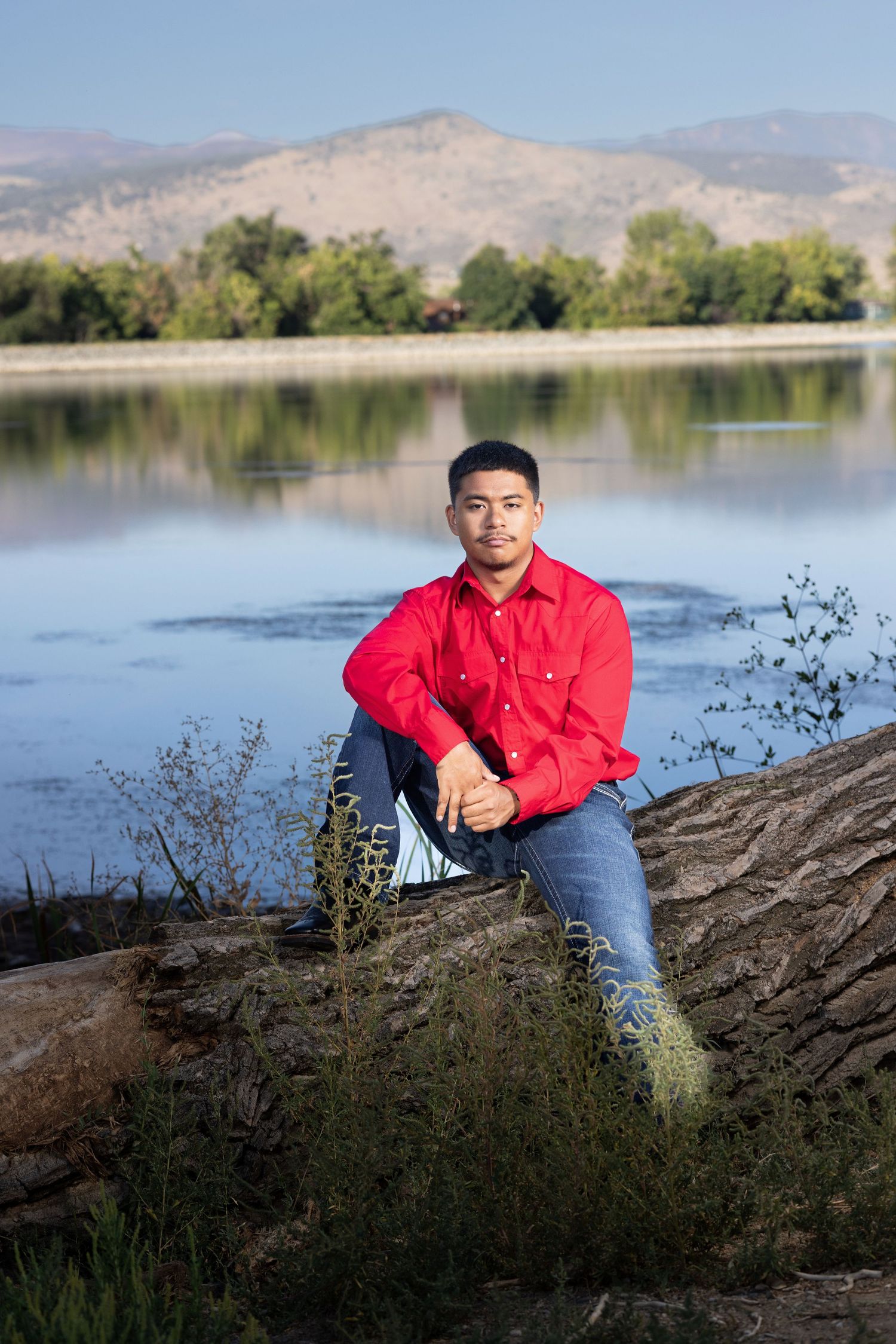 Young man in red shirt sitting on a log by a calm lake with mountain and tree reflections in background.