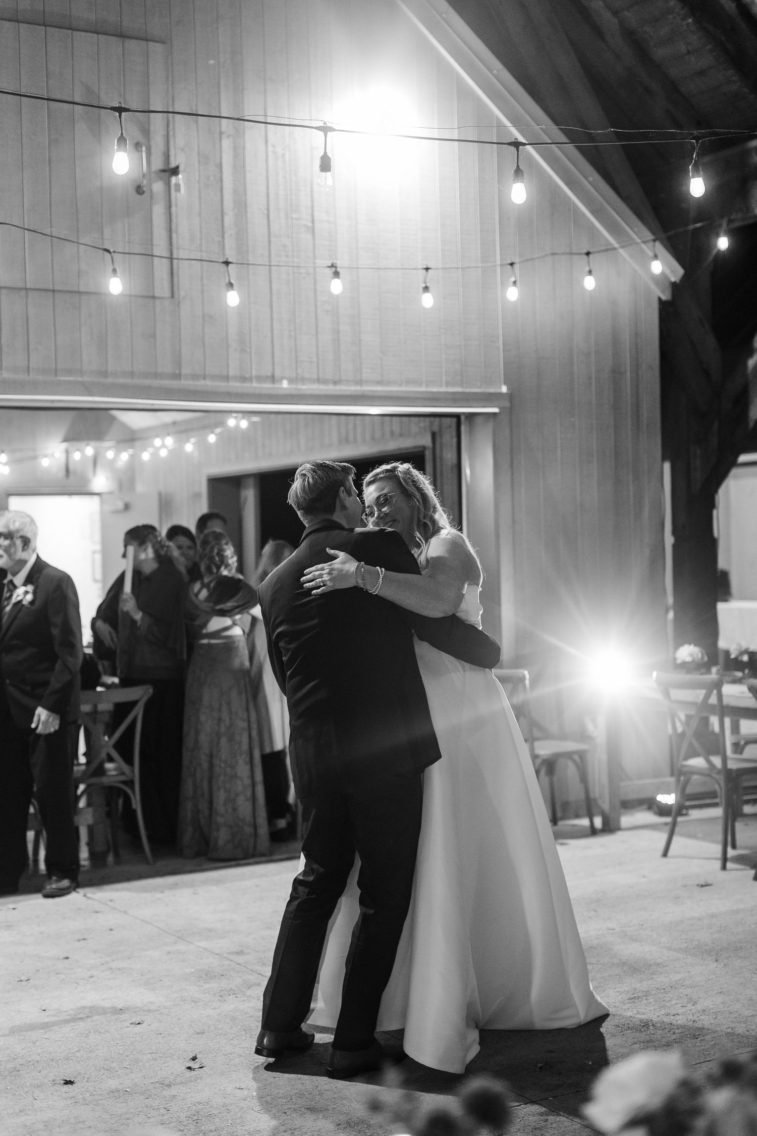 Bride in a white gown and groom in a dark suit share a first dance outside a barn under string lights as wedding guests watch.