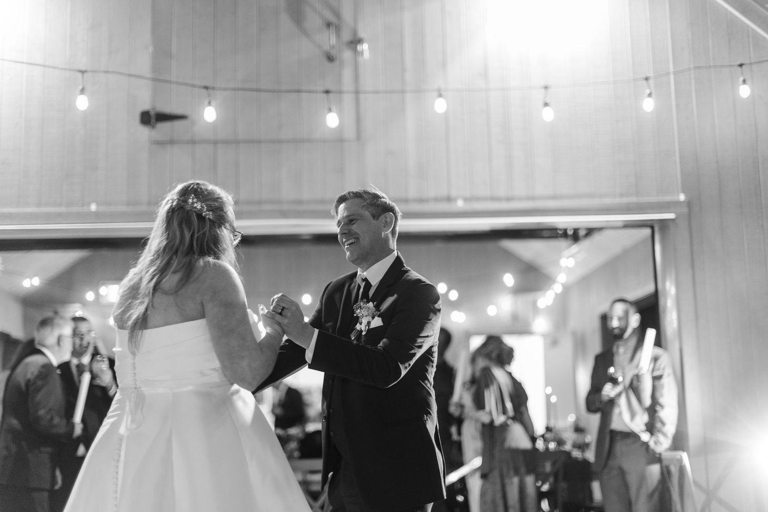 Black and white photo of a bride in a white gown dancing with a man in a suit at a wedding reception under string lights while guests watch.