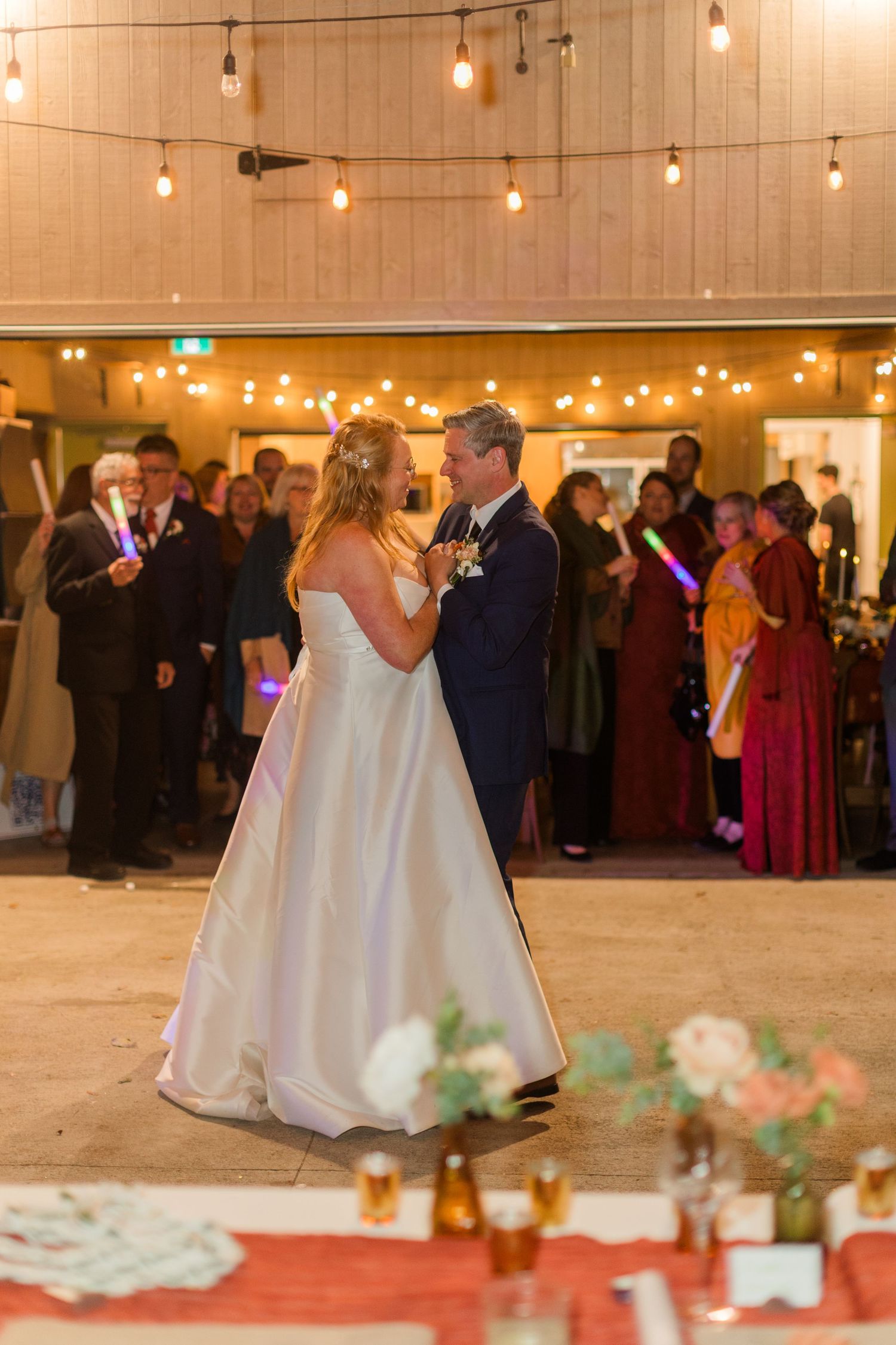 Bride in a white gown and groom in a navy suit share a first dance as guests with glow sticks watch under warm string lights.