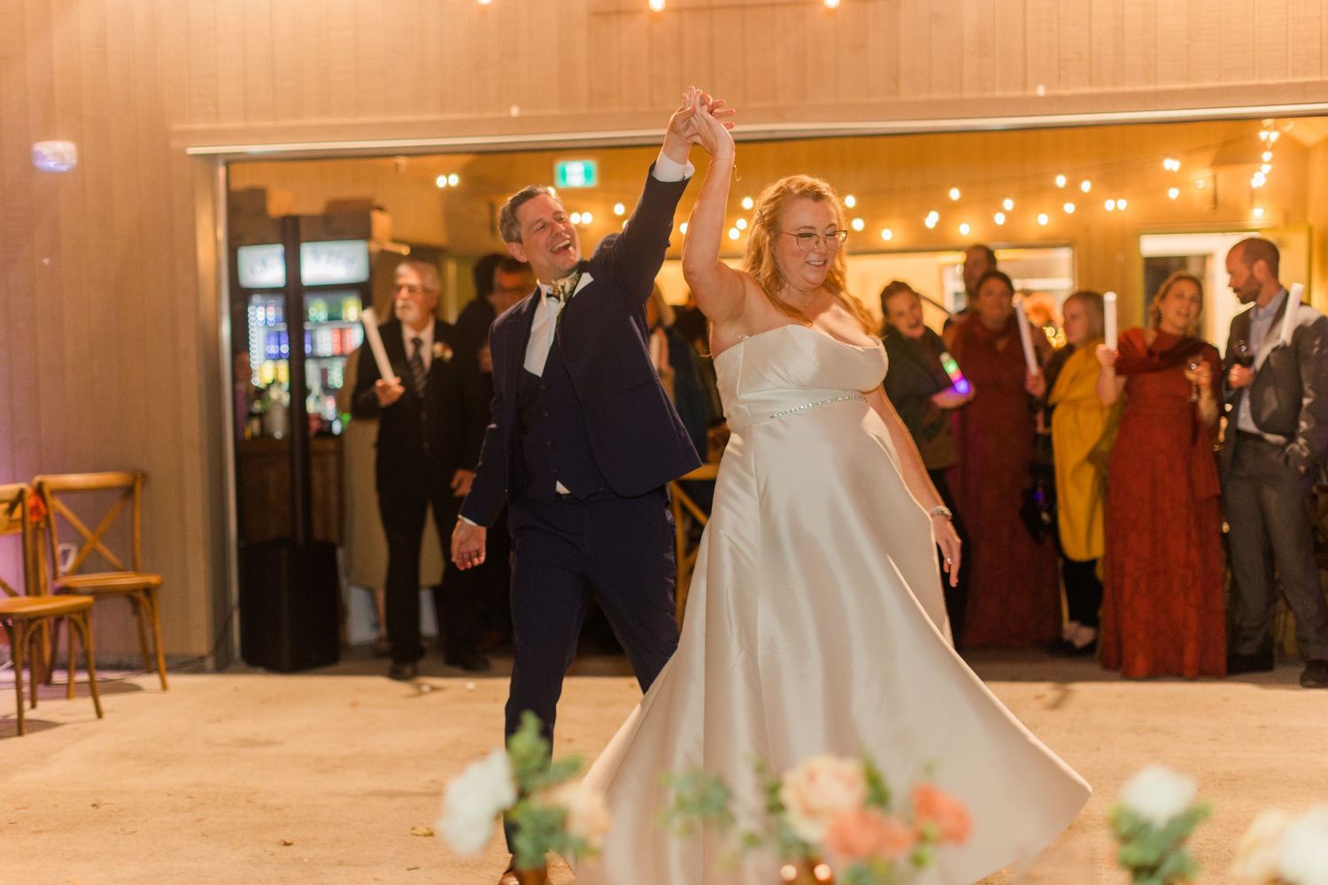 Bride in a white gown twirls on the dance floor with her groom in a navy suit as wedding guests watch and smile.