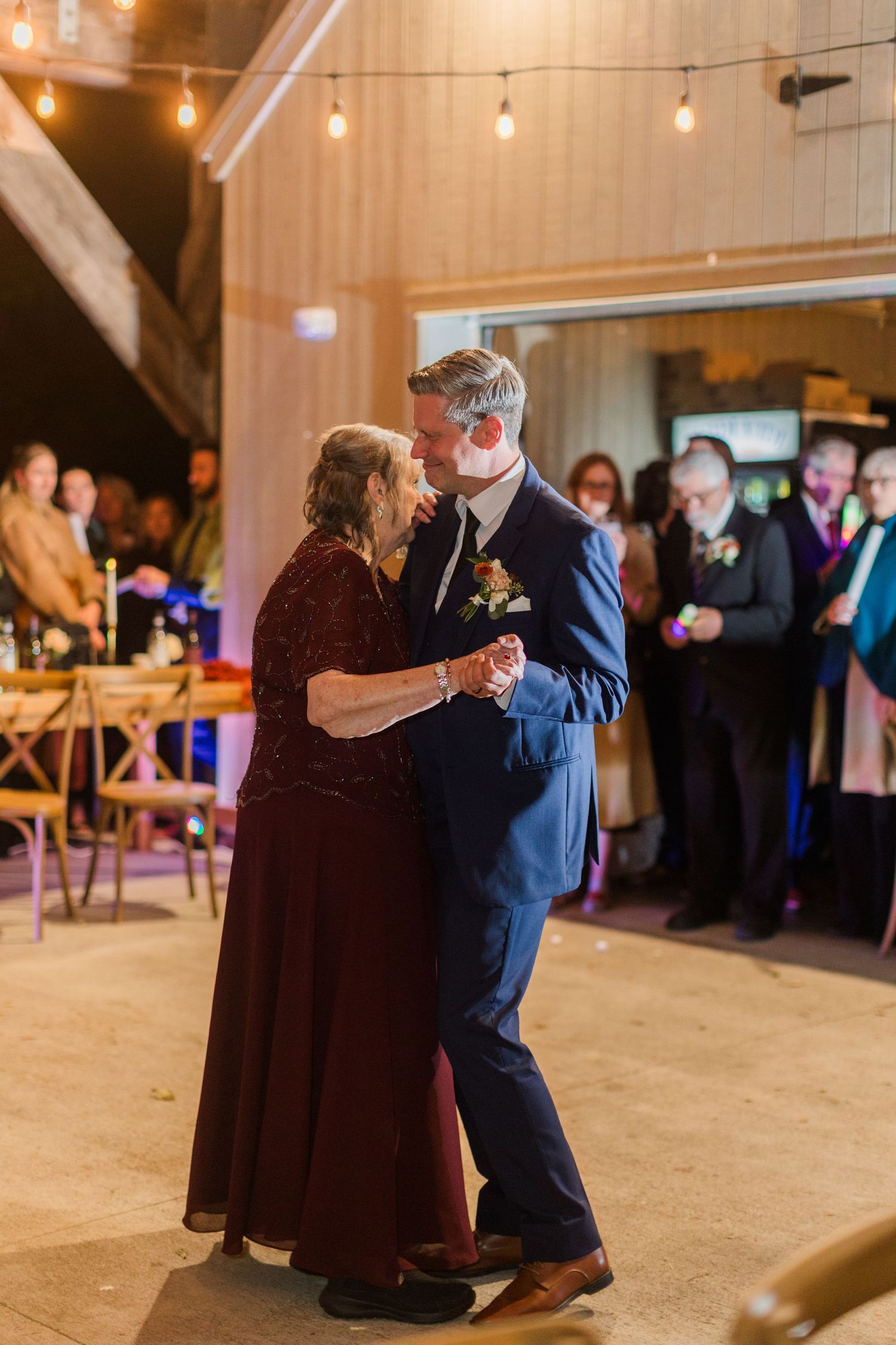 A groom in a navy suit dances with an older woman in a burgundy gown at a rustic barn wedding while guests watch under string lights.