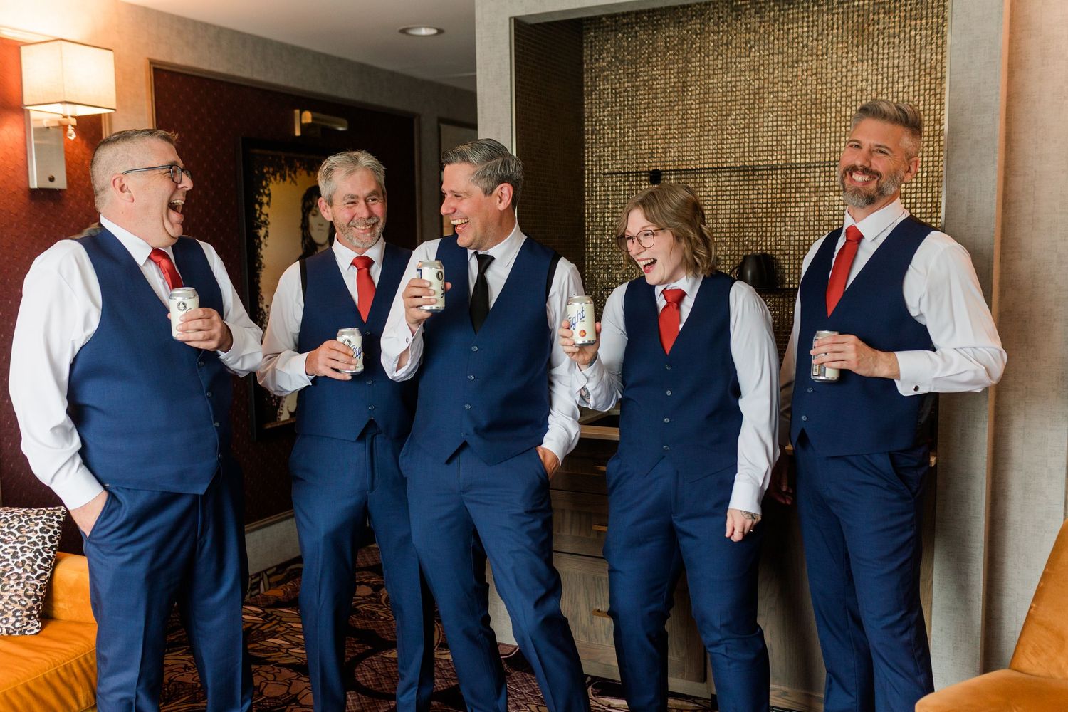 Five groomsmen in navy vests, white shirts and red ties laugh and hold drinks while getting ready together in a cozy hotel room.