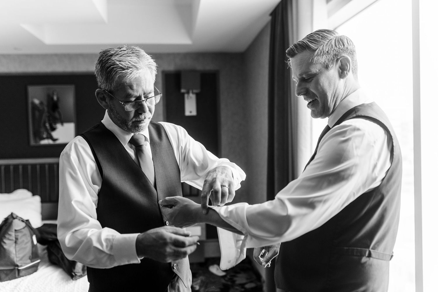 Two men in vests and dress shirts stand in a hotel room as one helps the other fasten his cufflink in a black and white photo.