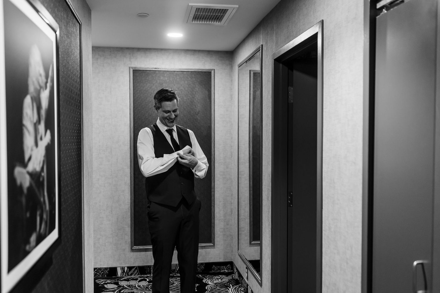 Black and white photo of a groom in a vest and tie adjusting his cuff while standing in a hotel hallway with framed art.