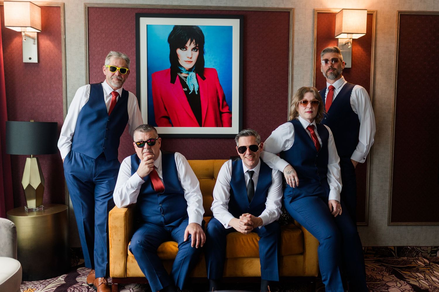 Five men in matching navy vests, white shirts, red ties and sunglasses pose on a gold sofa beneath a framed portrait in a stylish room.