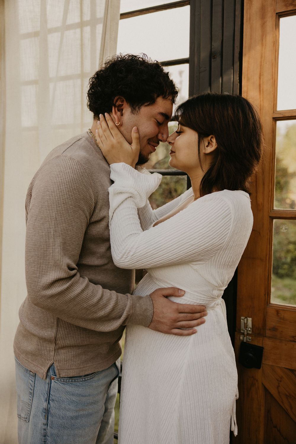 A couple stands forehead to forehead in a tender embrace beside a wooden door, the woman in a white dress and the man in a beige sweater.