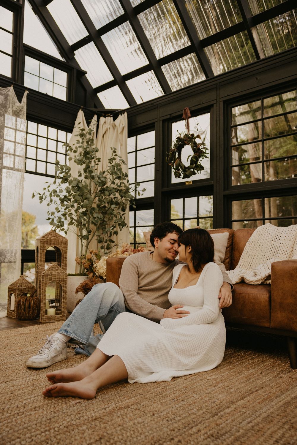Expecting couple in a white dress and tan sweater embracing on a jute rug in a sunlit glass greenhouse with a leather sofa.
