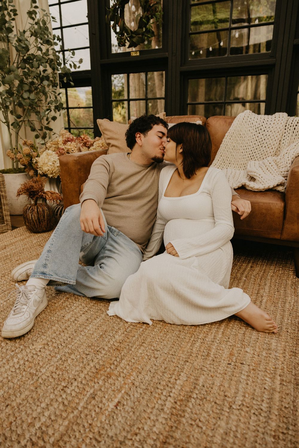 Couple kissing while seated on a woven jute rug beside a leather sofa in a bright sunroom, maternity-style photo shoot.