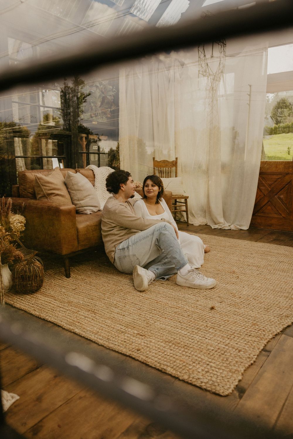 Couple sitting close together on a woven jute rug in a bright, rustic glass room with a leather sofa, sheer curtains and boho decor.