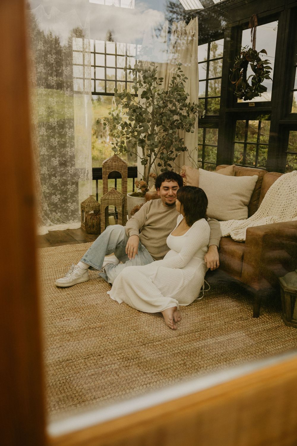 Couple sitting together on a woven jute rug in a sunlit greenhouse sunroom with a leather sofa, potted plant, and wreath.