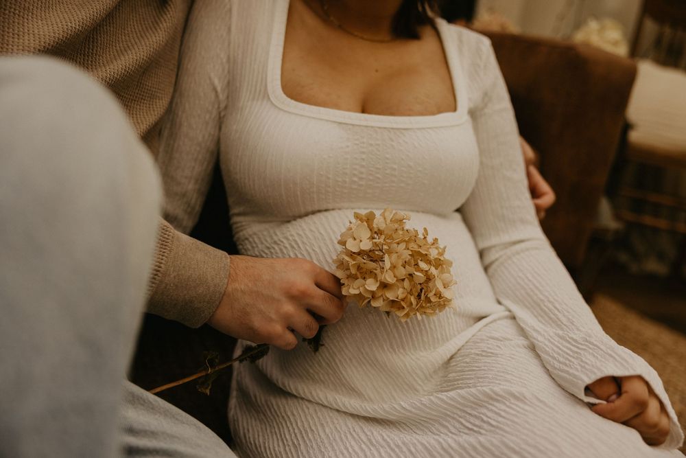 Pregnant woman in a white ribbed knit dress holding a dried hydrangea flower while another person's hand rests gently on her baby bump.