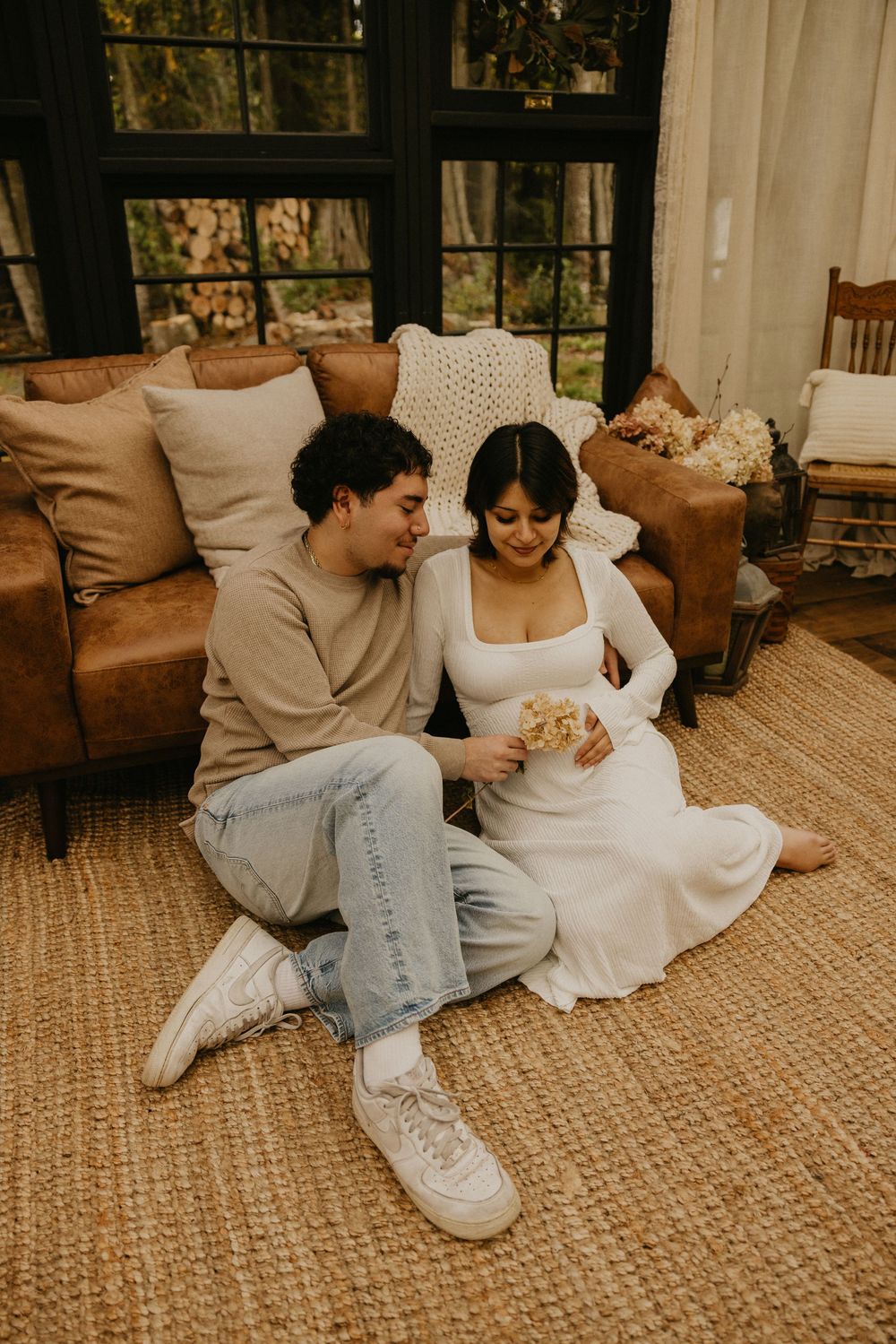 Smiling couple sitting on a woven jute rug in a sunlit room, the woman in a white dress holding dried flowers beside a brown leather sofa.