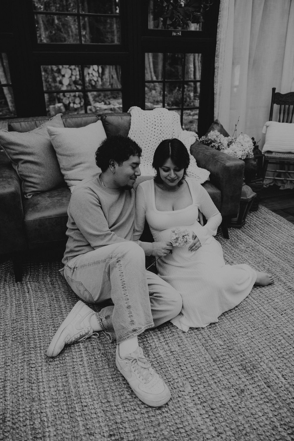 Black and white photo of a couple sitting close together on a woven rug in front of a sofa, smiling softly in a cozy sunlit room.