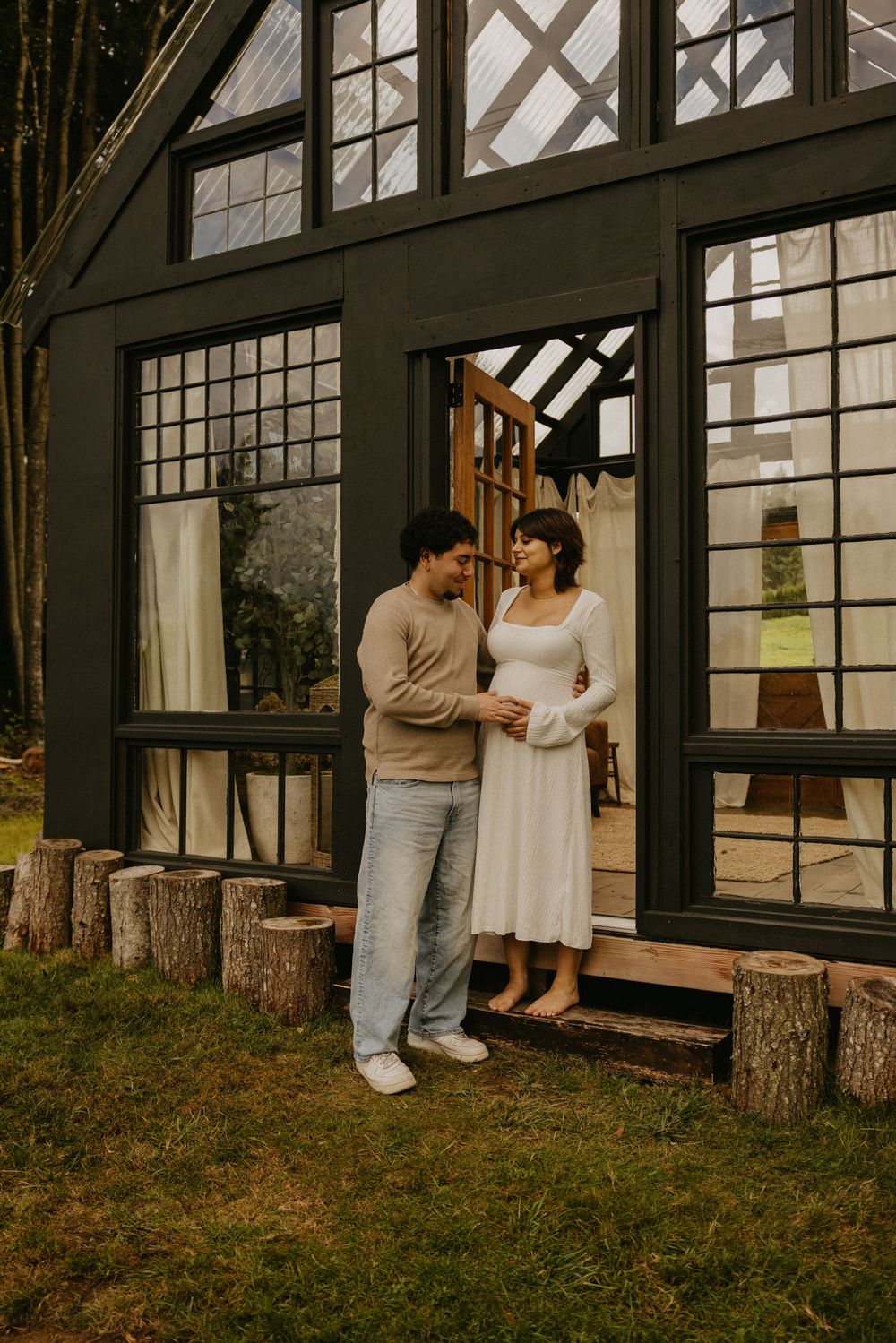 Expecting couple embracing in the doorway of a dark modern glass cabin surrounded by tree stump seating and green grass.