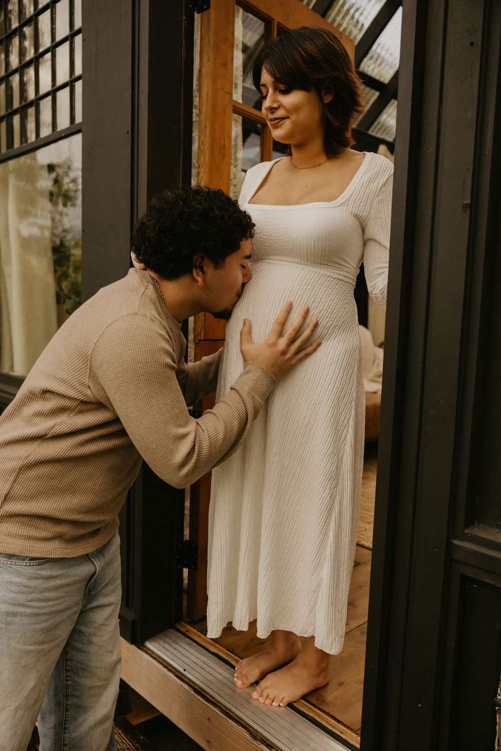 A man in a tan sweater kisses the belly of his pregnant partner, who stands barefoot in a white dress in a glass-walled doorway.