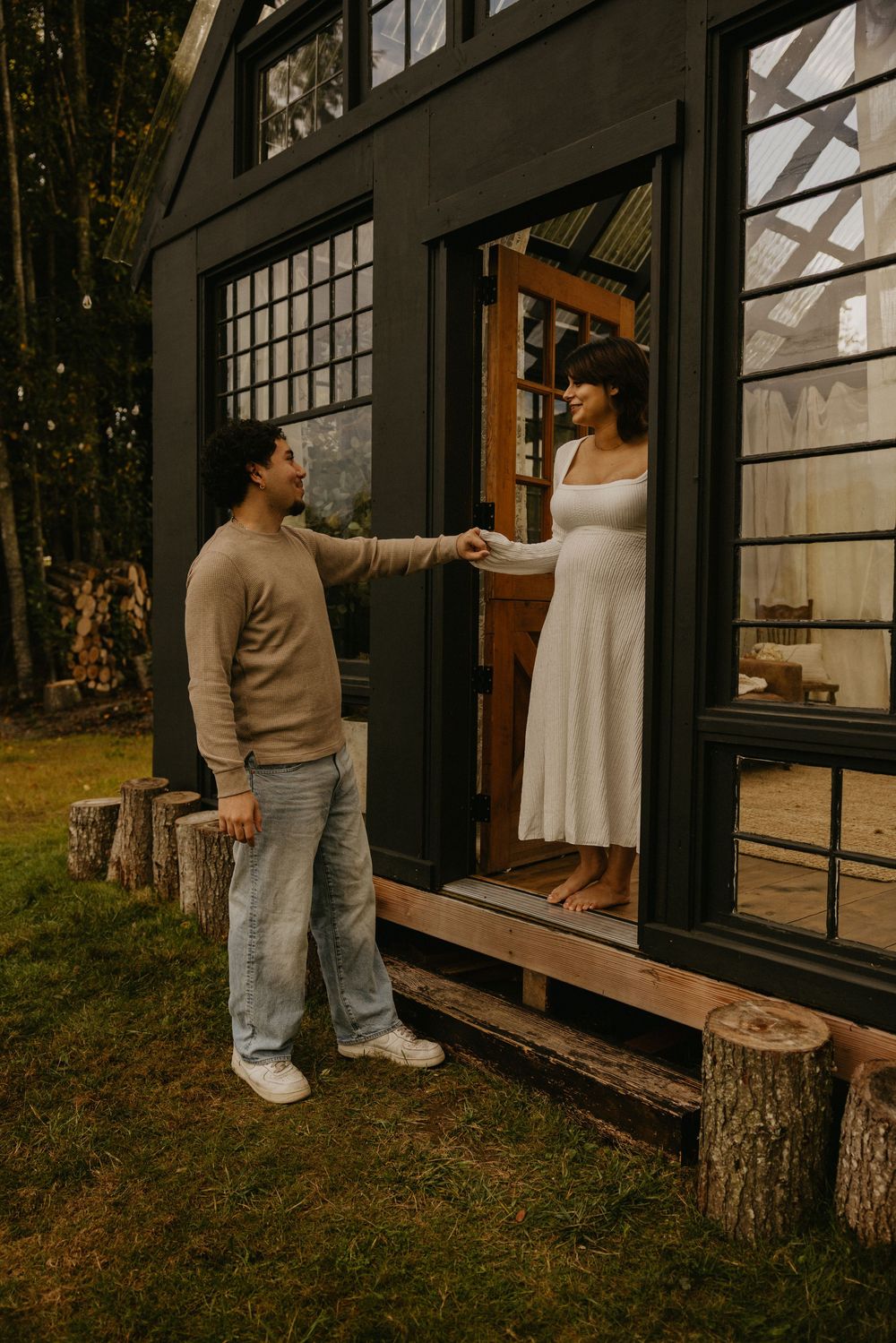 A man in a beige sweater and jeans holds hands with a woman in a white dress standing in the doorway of a black tiny house in a forest.