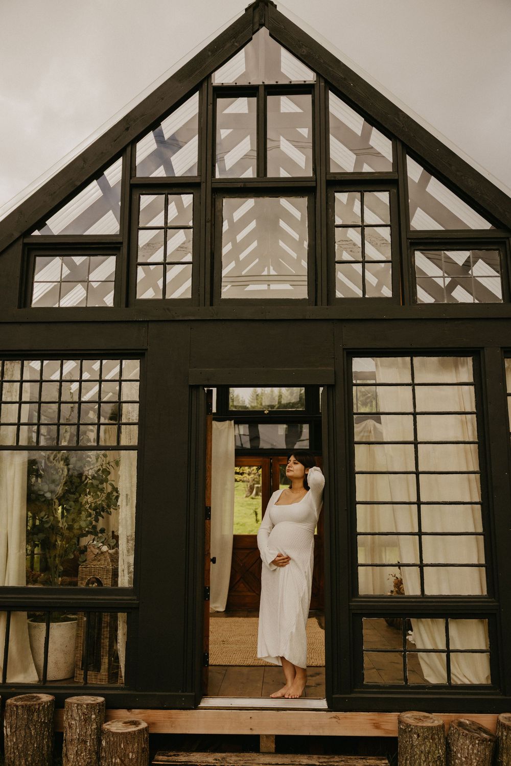 Woman in a flowing white dress stands barefoot in the doorway of a black-framed glass cabin with a peaked roof and log steps.