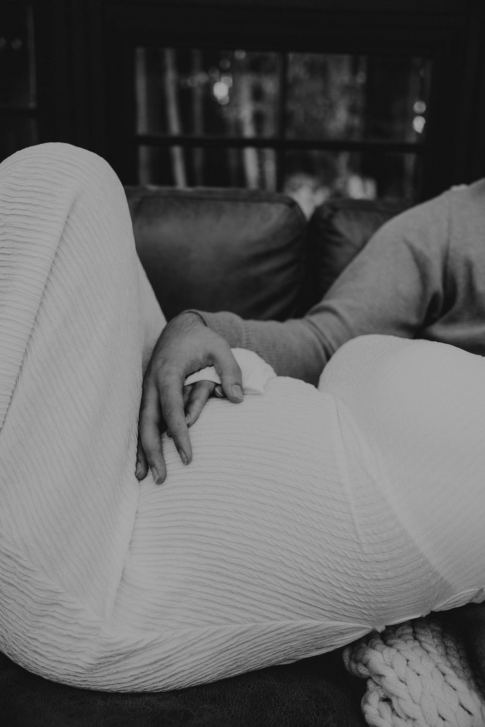 Black and white photo of a woman in a ribbed knit outfit reclining on a leather couch with her hand resting gently on her belly.