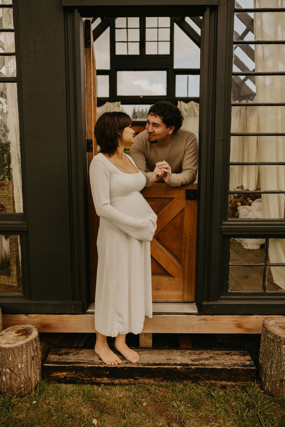 Pregnant woman in a white dress holds hands with her partner leaning from a rustic wooden barn-style door of a dark-framed glass cabin.