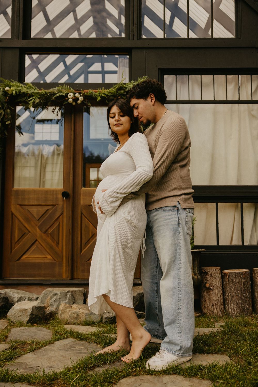 Expecting couple embracing in a maternity photo shoot, the pregnant woman in a white dress barefoot beside a rustic wooden greenhouse door.