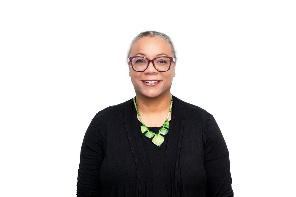 Smiling woman with gray hair, green geometric necklace, and red glasses wearing a black cardigan on white background.