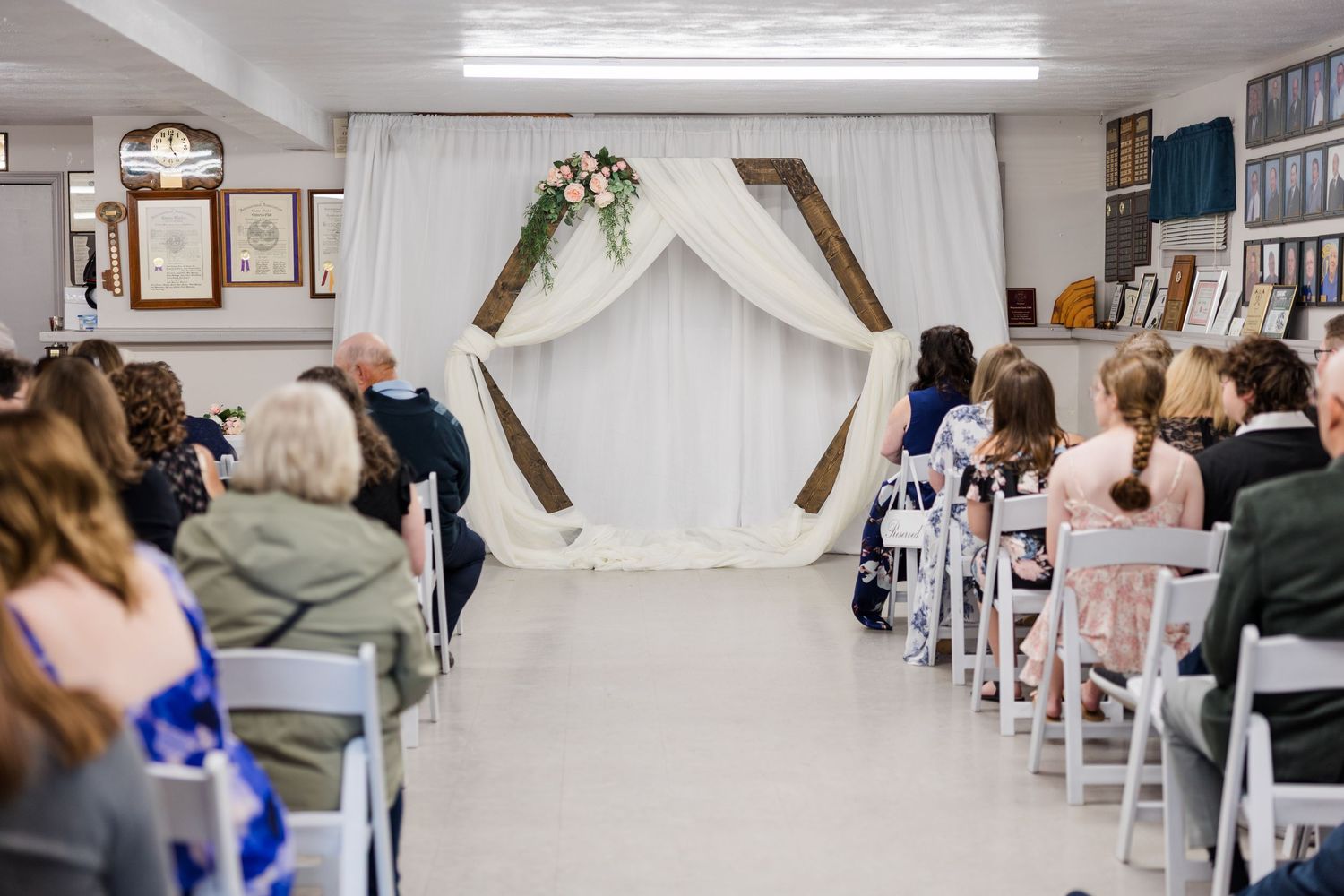 Indoor wedding ceremony with wooden arch, white draping, and floral decor, guests seated in white chairs facing the altar.