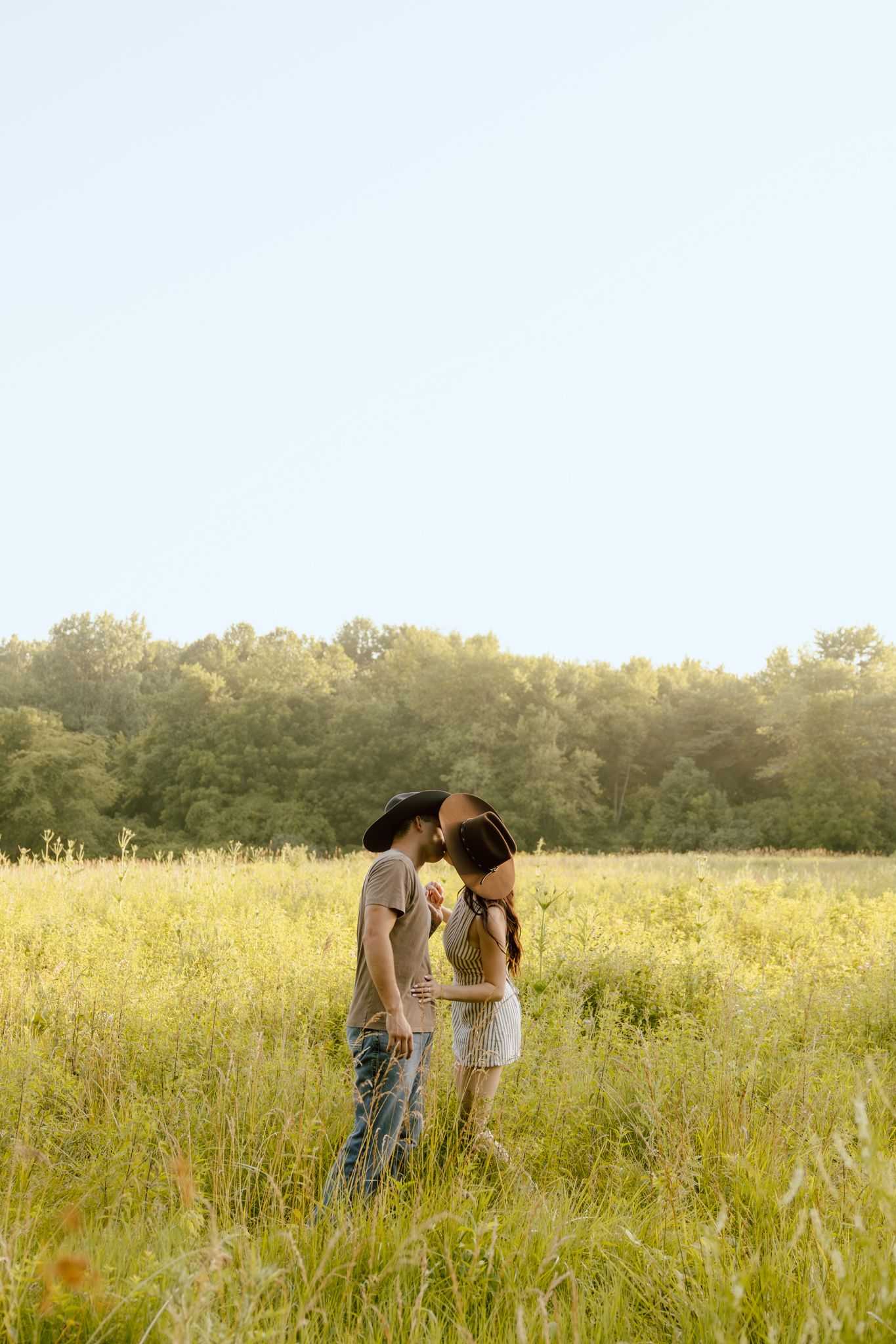 Couple kissing in a sunny meadow with tall grass and green trees in the background on a clear day.