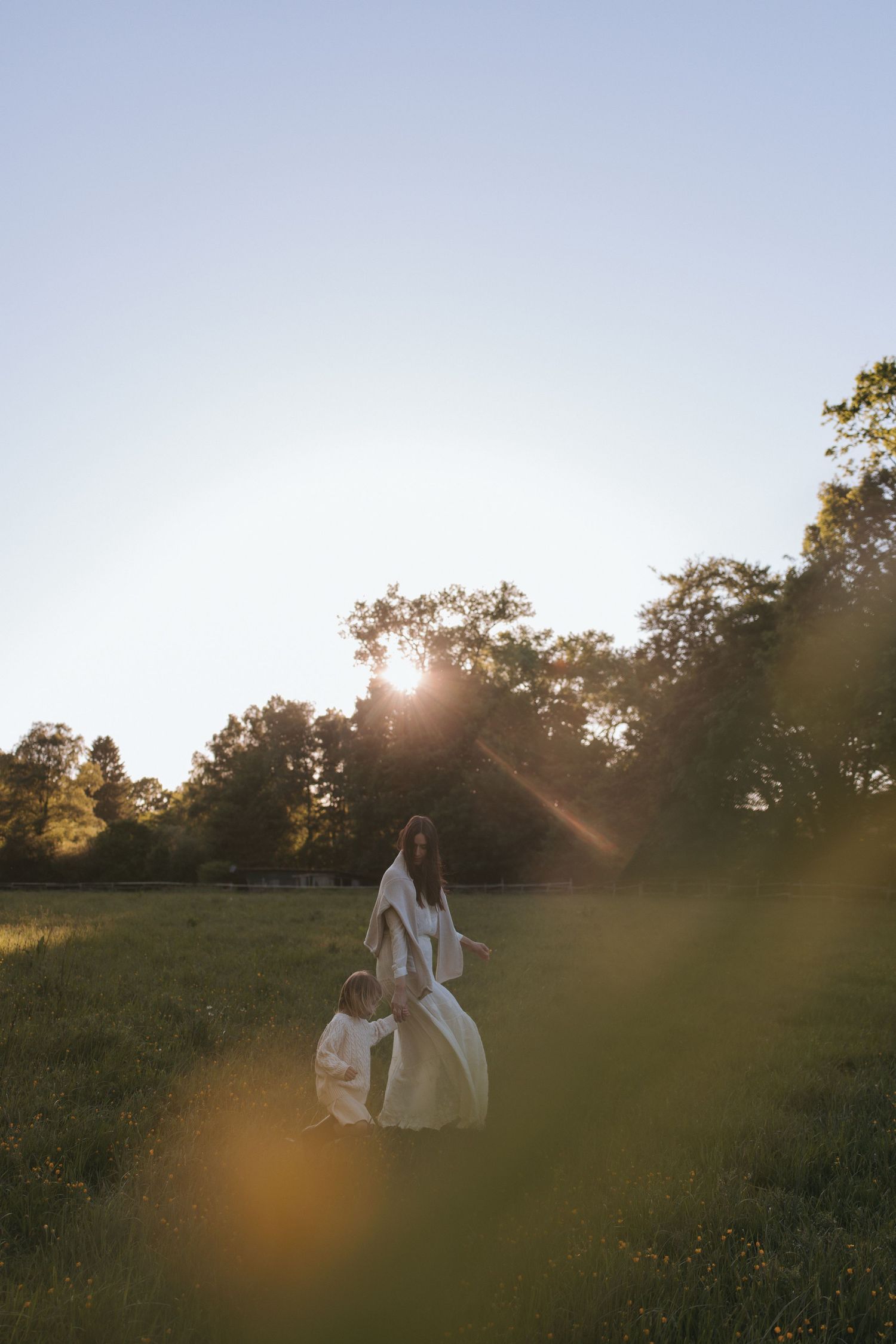 Mother and child in white dresses walking through a sunlit green meadow with trees in the golden hour light.