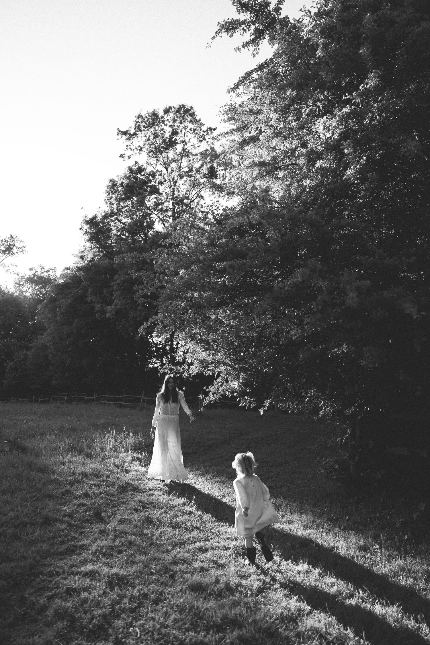 Black and white photo of a bride in white dress walking with a child through a sunlit grassy field near trees in New Zealand.