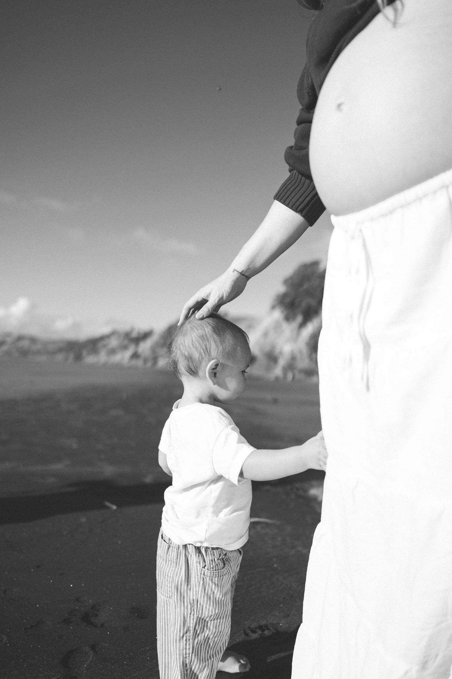 Black and white photo of a pregnant woman tenderly touching her toddler's head on a beach in Raglan New Zealand.