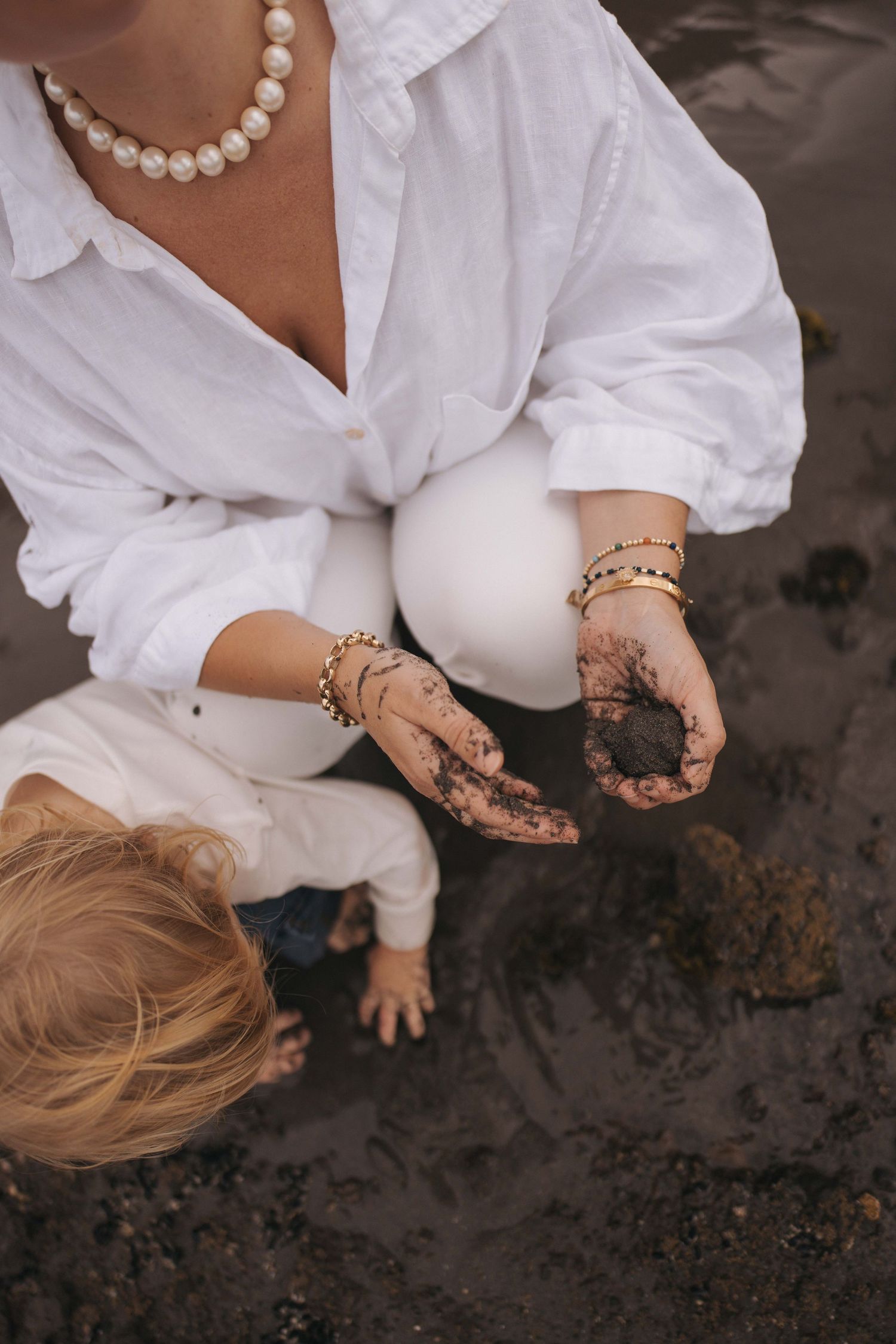 Woman in white shirt and pearl necklace crouching on beach, showing muddy hands to a young toddler beside her.