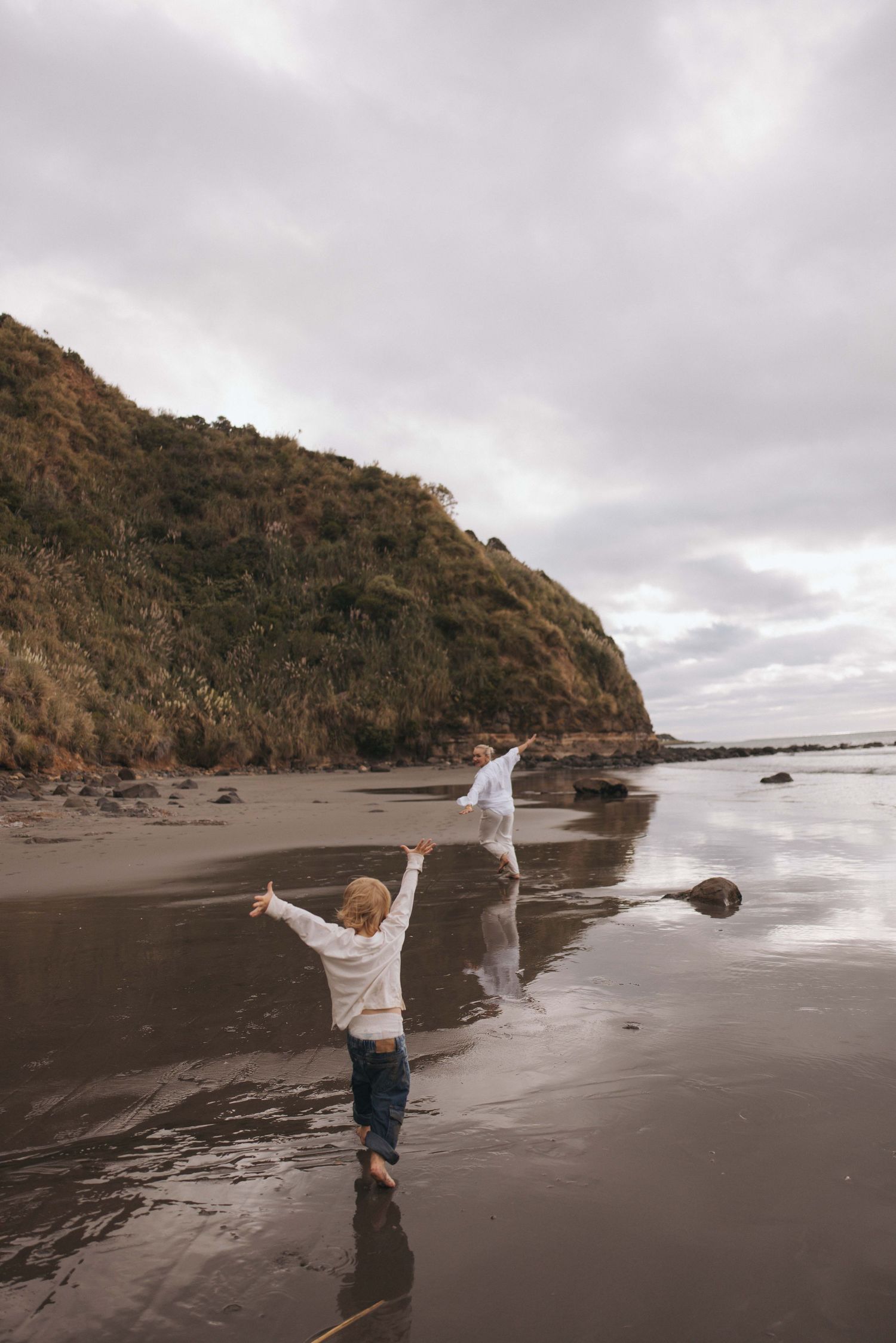 Two people joyfully running on a dark sandy beach with arms outstretched, with a green hillside and cloudy sky backdrop.