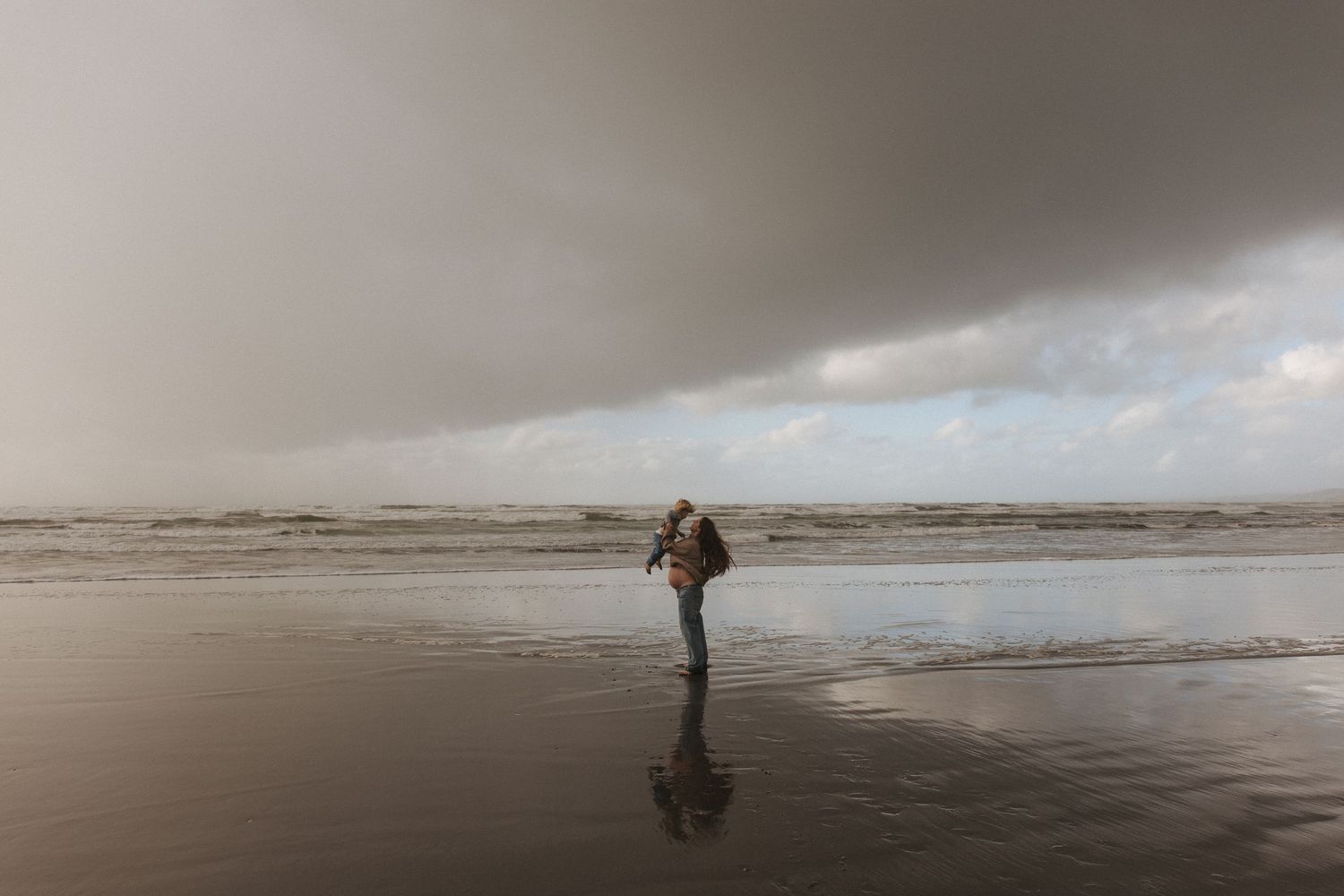 A pregnant mother stands on a reflective wet beach under dramatic stormy skies near the ocean shoreline in Raglan New Zealand.