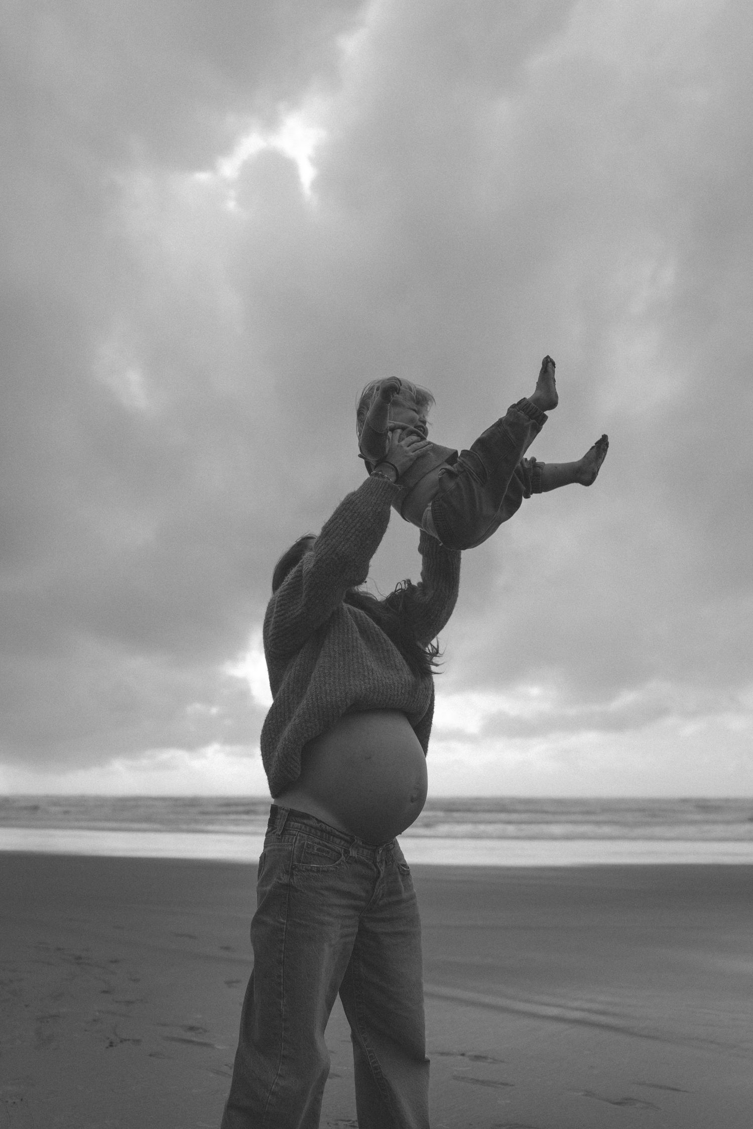 Black and white photo of parent lifting child overhead on a cloudy beach, waves visible in the background in Waikato.