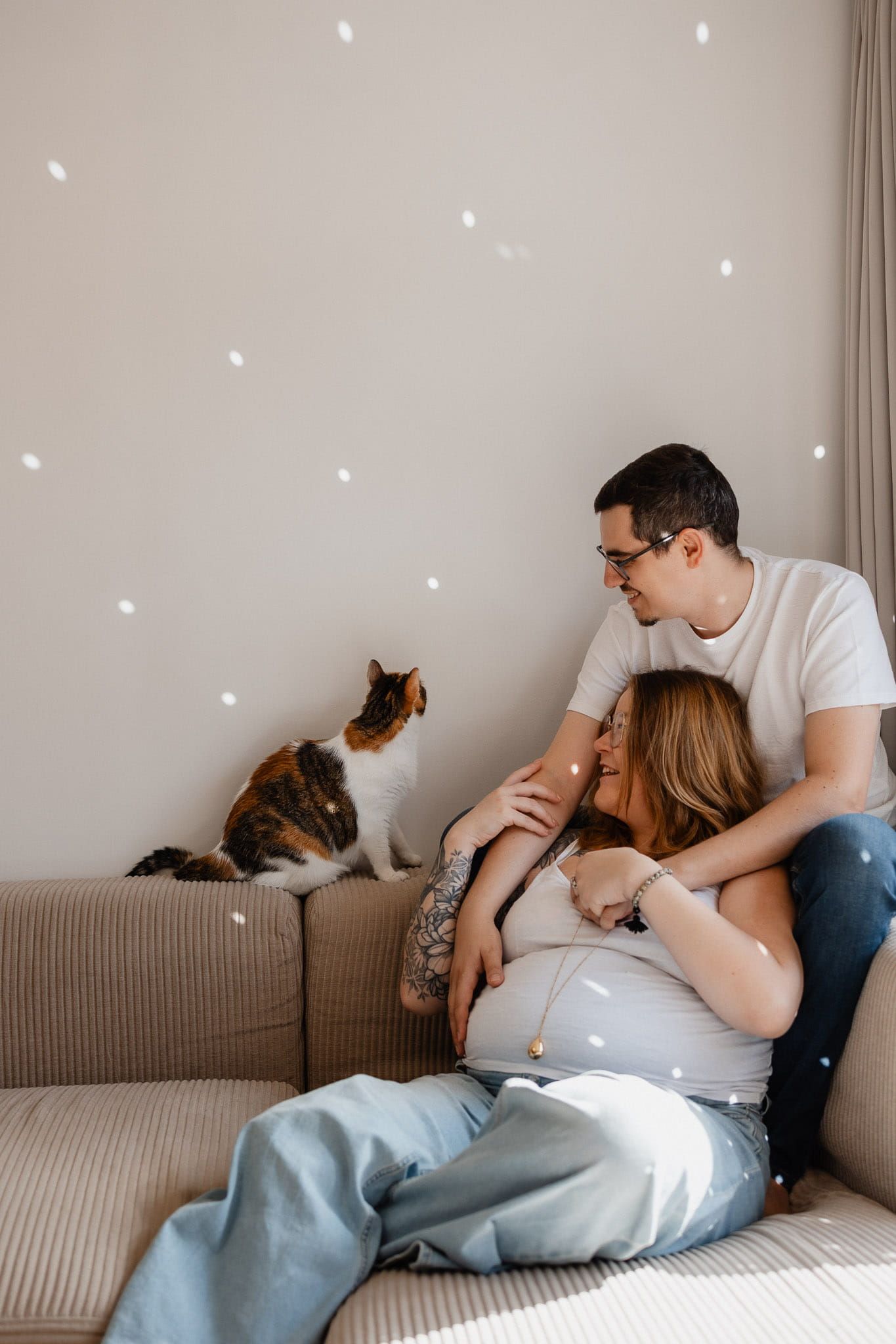 Femme enceinte et son compagnon rient car le chat essaie d'attraper les points lumineux de la boule a facette pendant leur seance photo